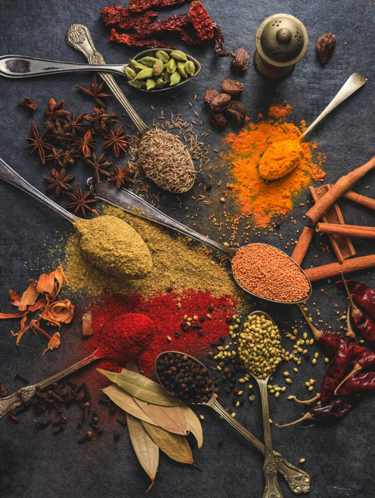 A vibrant assortment of herbs and spices, including turmeric, cumin, cardamom, cinnamon, chili, star anise, and black pepper, arranged on dark textured background with vintage spoons