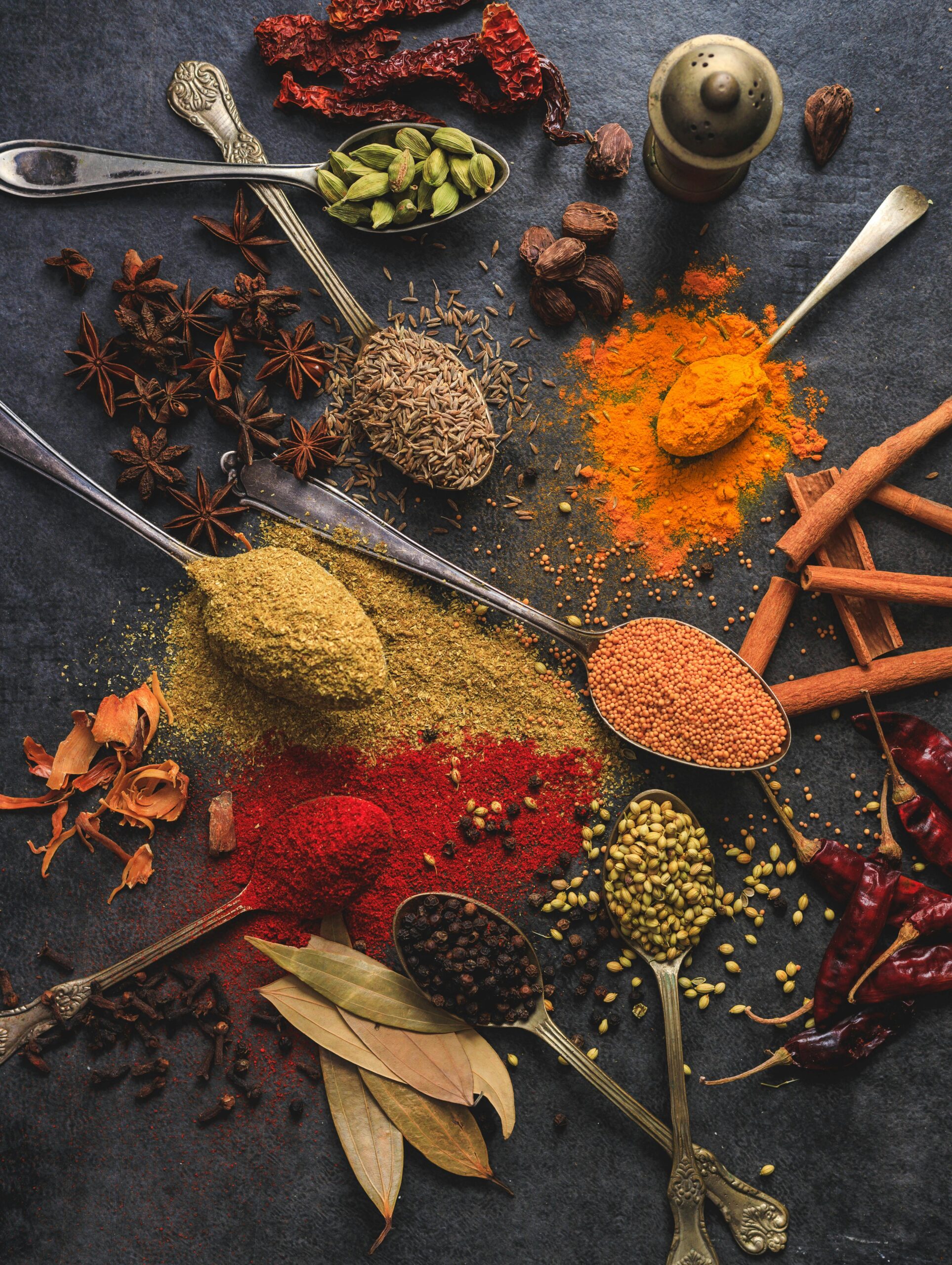 A vibrant assortment of herbs and spices, including turmeric, cumin, cardamom, cinnamon, chili, star anise, and black pepper, arranged on dark textured background with vintage spoons