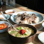 Bowl of Korean beef soup (Seolleongtang) topped with green onions and red chili paste, served with sliced beef and kimchi on the side.