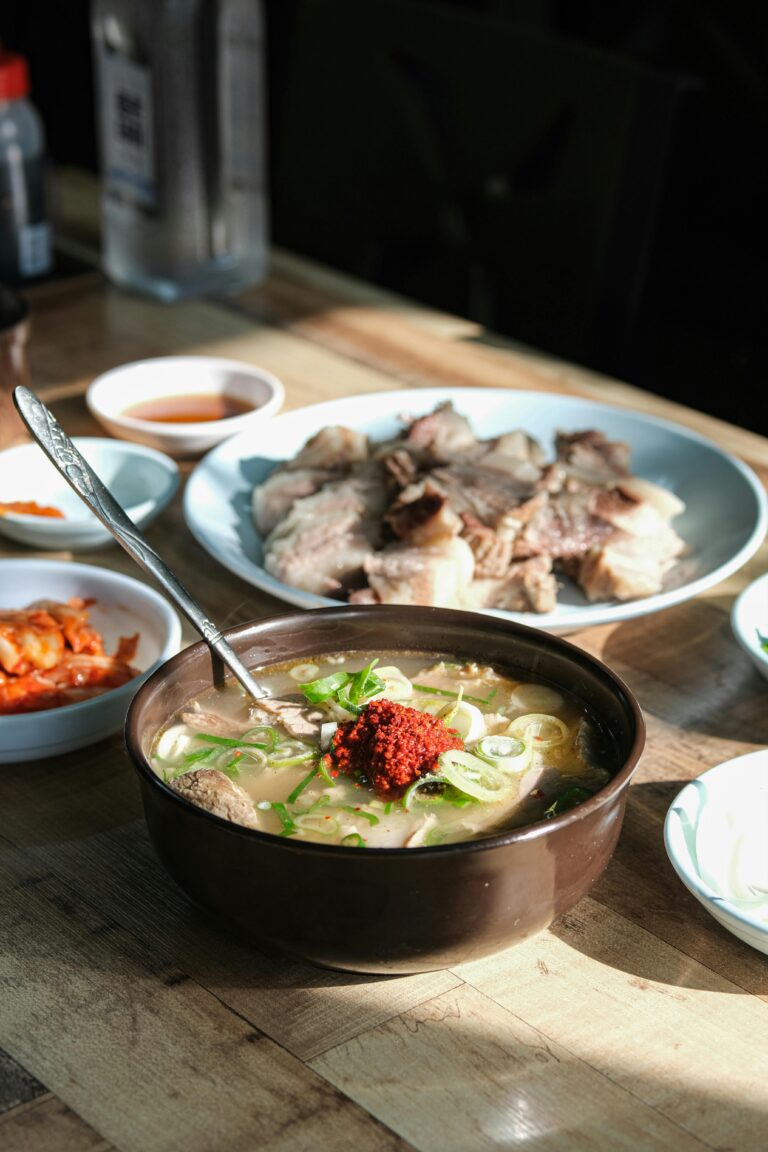 Bowl of Korean beef soup (Seolleongtang) topped with green onions and red chili paste, served with sliced beef and kimchi on the side.