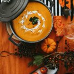 A festive pot of Spicy Korean Pumpkin Soup garnished with cream and parsley, surrounded by pumpkins and autumn-themed decor on an orange and striped background.